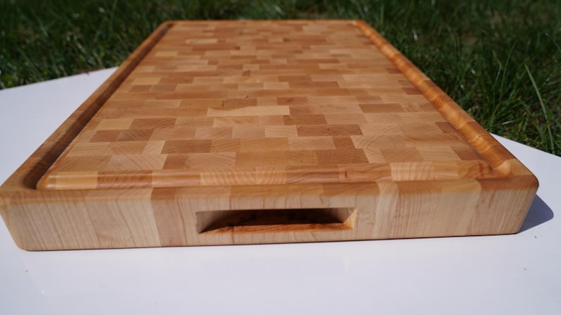 May include: A rectangular end-grain wooden cutting board featuring a light tan and honey-colored mosaic pattern. The board includes a carved juice groove along the perimeter and a recessed handle cutout on the front edge for easy lifting.