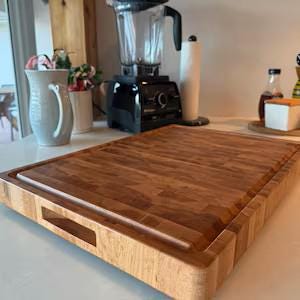 May include: A rectangular wooden cutting board with a thick, multi-layered grain pattern and a carved handle slot on the front edge. The board features a recessed juice groove along the perimeter and a smooth, polished surface finish.