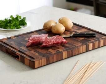 May include: A large wooden cutting board with a checkered pattern in shades of brown. Two raw steaks, a knife, and potatoes are on the board. A bunch of wooden skewers are in the foreground.