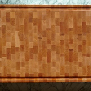May include: A rectangular wooden cutting board featuring an end-grain construction with a mosaic of light and medium brown wood blocks. The board has a recessed juice groove running along the perimeter of its top surface with rounded edges.