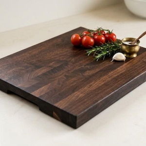 May include: A dark brown wooden chopping board with a rectangular shape and visible wood grain. It is adorned with red tomatoes, rosemary, a garlic clove, and a small brass bowl with a spoon.