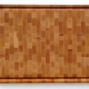 May include: A rectangular wooden cutting board featuring a mosaic-like end-grain pattern composed of various light and medium brown wood blocks. The board has a smooth surface and a recessed juice groove running along the entire perimeter edge.