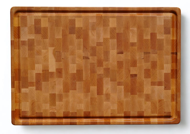 May include: A rectangular wooden cutting board featuring a mosaic-like end-grain pattern composed of various light and medium brown wood blocks. The board has a smooth surface and a recessed juice groove running along the entire perimeter edge.