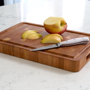May include: A rectangular wooden cutting board with a light brown finish. Sliced apple pieces and a partially cut apple rest on the board. A small knife with a white handle is also on the board.