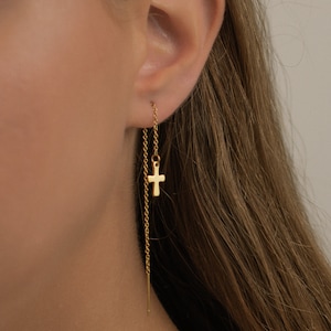 Peut inclure: Boucles d'oreilles en chaîne dorée avec un petit pendentif en forme de croix. Les boucles d'oreilles sont enfilées dans le lobe de l'oreille et pendent.
