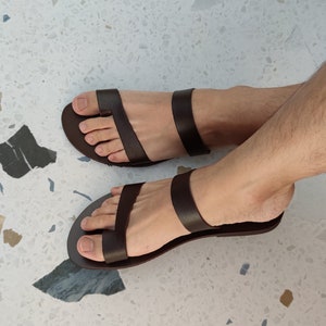 May include: A pair of brown leather sandals with a simple design. The sandals have a single strap that goes across the foot and a toe loop.