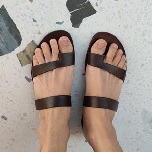 May include: A pair of brown leather sandals with two straps across the foot. The sandals are being worn on a person's feet.