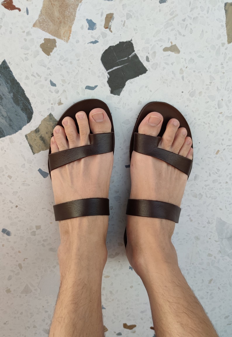May include: A pair of brown leather sandals with two straps across the foot. The sandals are being worn on a person's feet.
