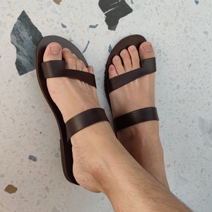 May include: A pair of brown leather sandals with two straps across the foot. The sandals have a toe loop and are being worn.