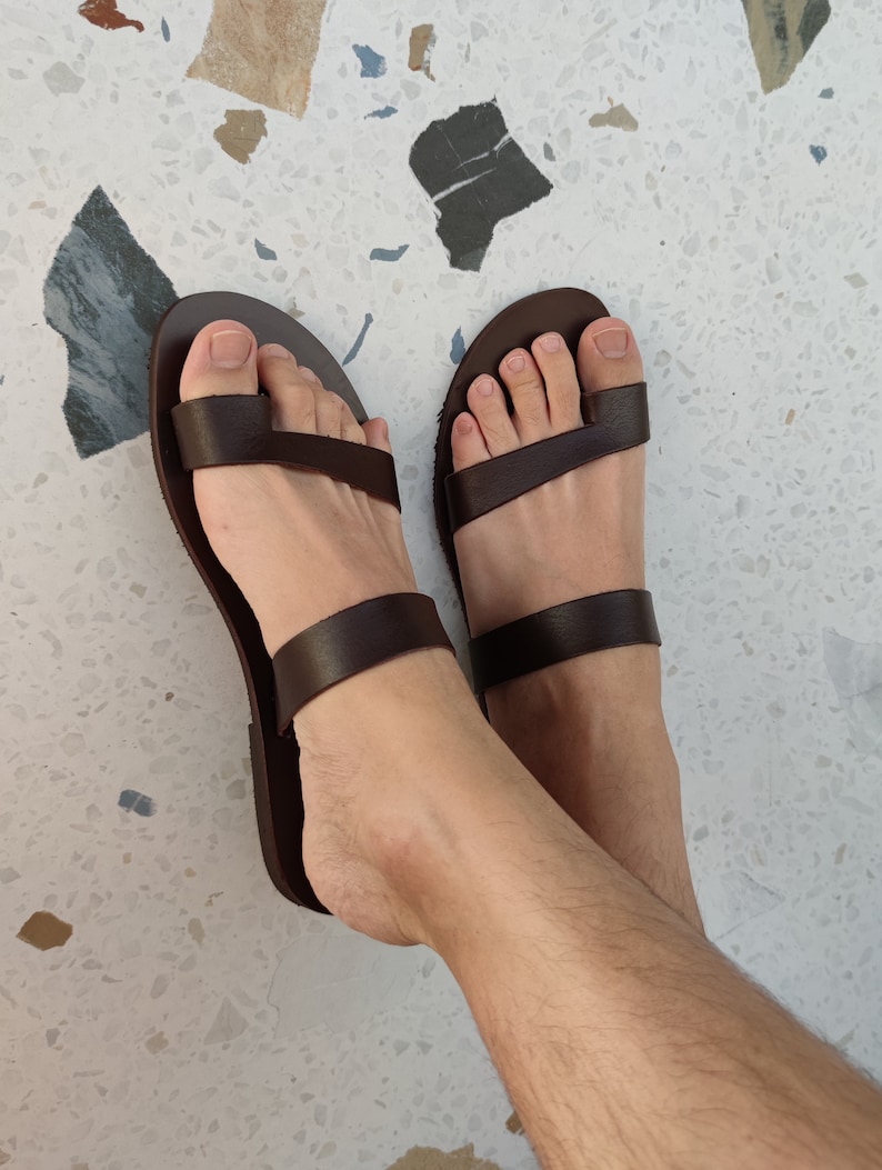 May include: A pair of brown leather sandals with two straps across the foot. The sandals have a toe loop and are being worn.