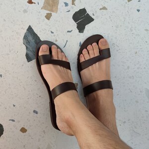 May include: A pair of brown leather sandals with two straps across the foot. The sandals are being worn on a person's feet.