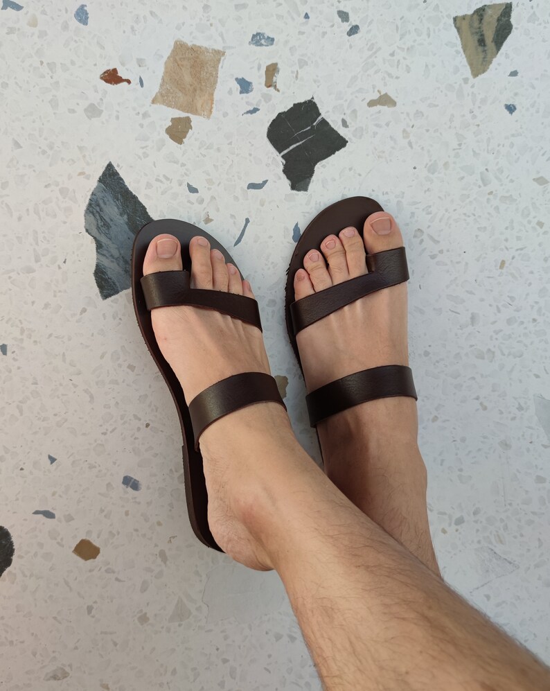 May include: A pair of brown leather sandals with two straps across the foot. The sandals are being worn on a person's feet.