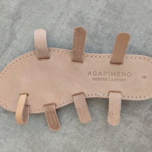 May include: The underside of a light brown leather sandal with multiple straps. The sandal is labelled "AGAPIMENO GENUINE LEATHER".