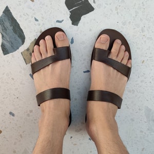 May include: A pair of brown leather sandals with two straps across the foot. The sandals are being worn on a person's feet.