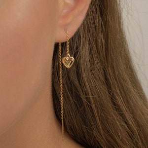 Puede incluir: Pendiente de cadena de oro con un dije de corazón de oro. La cadena es larga y cuelga por debajo del lóbulo de la oreja.