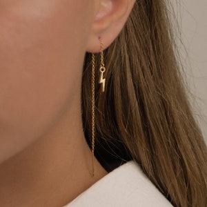 May include: Gold chain earrings with a small lightning bolt charm hanging from the bottom. The earrings are worn in a person's ear.
