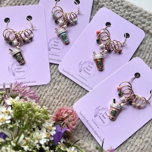 Könnte beinhalten: Vier Sets Maschenmarkierer mit goldfarbenen Ringen, Perlen und Miniatur-Kaffeebecher-Anhängern in verschiedenen Farben. Jedes Set ist an einer lavendelfarbenen Karte mit dem Text "Hand Dyed Harmony" und einer E-Mail-Adresse befestigt.