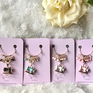 Pode incluir: Quatro cartões cor-de-rosa com anéis dourados e um amuleto de máquina de costura. Cada cartão tem o texto "Hand Dyed Harmony" e um endereço de site. Os amuletos são preto, turquesa, branco e rosa.
