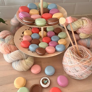 Peut inclure: Un présentoir en bois à deux étages avec des contenants en forme de macarons de couleur pastel. Le présentoir est entouré de pelotes de laine dans différentes nuances de rose, bleu et jaune. Il y a aussi plusieurs contenants de macarons sur la table, certains ouverts et d'autres fermés.