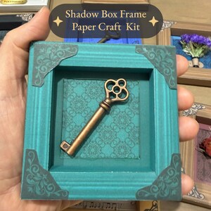 May include: A teal shadow box frame paper craft kit featuring a bronze-colored skeleton key mounted on a teal background with a repeating geometric pattern. The frame has dark decorative corner accents and the text Shadow Box Frame Paper Craft Kit.