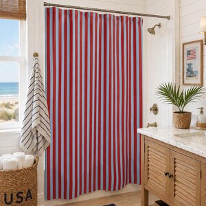Könnte beinhalten: Ein rot und hellblau gestreifter Duschvorhang hängt in einem Badezimmer. Ein Weidenkorb mit dem Wort "USA" steht auf dem Boden. Ein Fenster mit Blick auf das Meer ist sichtbar.