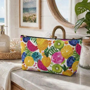 Può includere: Un beauty case rettangolare con un vivace motivo a frutta. Il design presenta illustrazioni colorate di anguria, kiwi, arance e altri frutti tropicali su sfondo bianco. La borsa ha una cerniera nera.