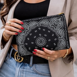 May include: Black clutch with a white paisley design, held by a person wearing a tan blazer, black top, and blue jeans. The clutch has a gold zipper. Gold jewelry and a black belt with a gold buckle complete the look.