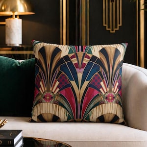 May include: A decorative throw pillow with an Art Deco design in shades of beige, black, red, blue, and gold. The pillow is on a cream-colored sofa, with a dark green pillow to the left. The background features gold accents and a lamp.