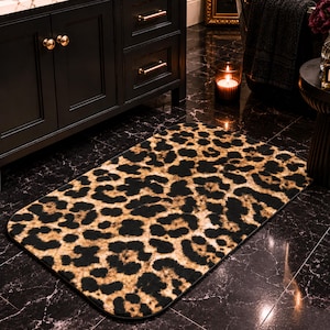 May include: Rectangular bath mat with a leopard print pattern in shades of brown and black. The mat has rounded corners and is placed on a black marble floor. The bathroom setting includes a black vanity, a candle, and a small side table.