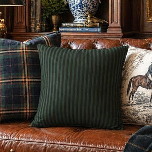 May include: A brown leather sofa featuring three decorative pillows: one with a dark green and blue plaid pattern, one with dark green vertical stripes, and one with a cream-colored fabric depicting a person riding a brown horse in a landscape.