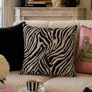 Pode incluir: Três almofadas quadradas num sofá creme: uma preta lisa, uma com padrão de riscas de zebra a preto e branco e uma almofada cor-de-rosa com uma ilustração gráfica de um leopardo num banco verde com rebordos pretos.