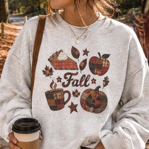 以下が含まれることがあります： Fallという文字、パイの切り身、チェック柄のリンゴ、チェック柄のマグカップ、チェック柄のカボチャが、秋の葉や星と共に描かれたグラフィックプリントのライトグレーのクルーネックトレーナー。ダークブルーのジーンズとサングラスを着用。