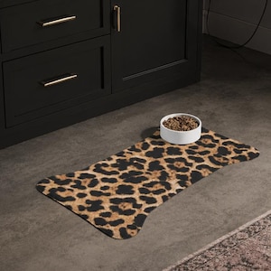 May include: A rectangular pet food mat with a leopard print pattern. A white ceramic bowl filled with pet food sits on the mat. The mat is on a dark gray floor in front of a black cabinet.