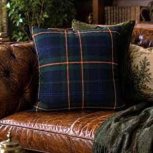 May include: A square throw pillow featuring a dark blue and green plaid pattern with thin red and yellow lines. The pillow is positioned on a brown tufted leather sofa alongside a dark green throw blanket and a decorative pillow with a tree print.