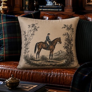 May include: A square throw pillow in a light beige fabric featuring a black ink-style illustration of a person riding a horse, framed by trees. The design is centered on the pillow, which is positioned on a brown leather sofa with other pillows.