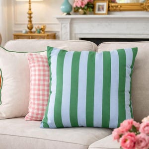 Könnte beinhalten: Gemütliches Wohnzimmer mit beigem Sofa und dekorativen Kissen. Ein Kissen mit grünen und hellblauen Streifen sticht hervor. Auf einem Couchtisch liegen Bücher und eine Tasse mit Herz. Eine Vase mit rosa Blumen sorgt für Farbtupfer.