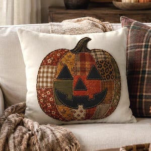 May include: A square off-white throw pillow featuring a central pumpkin applique. The pumpkin is constructed from various fabric patches in shades of orange, brown, green, and tan, with a black fabric jack-o'-lantern face stitched onto the front.