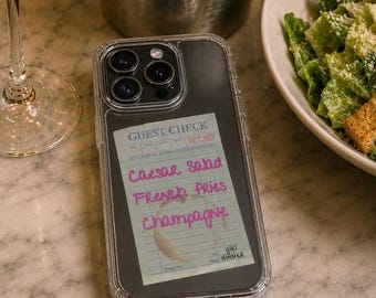 May include: A clear smartphone case featuring a guest check graphic with the text Caesar Salad, French Fries, and Champagne written in pink script, along with a circular stain mark, positioned on a table with a glass of champagne and food.