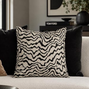 May include: A square throw pillow featuring a cream-colored base with a repeating black wavy, horizontal line pattern. The pillow is positioned on a light-colored sofa between a solid black throw pillow and a solid tan-colored throw pillow.