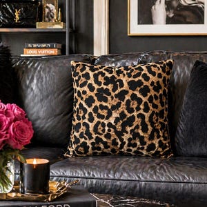 May include: A black leather sofa with a leopard print pillow and a black velvet pillow. A bouquet of pink roses sits on a gold tray with a candle and books. Artwork and a shelf with decor are in the background.