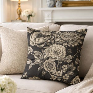 Może przedstawiać: Dwie dekoracyjne poduszki na beżowej sofie. Jedna poduszka ma kwiatowy wzór w kolorze czarnym i kremowym, druga ma kremowy, wzorzysty design. Kremowy koc jest zarzucony na ramię sofy.