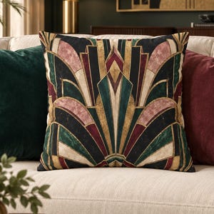 May include: A square throw pillow featuring a symmetrical geometric pattern in black, gold, deep green, and dusty pink. The design consists of sharp, angular lines and fan-like shapes radiating from the center, creating a bold, structured visual effect.