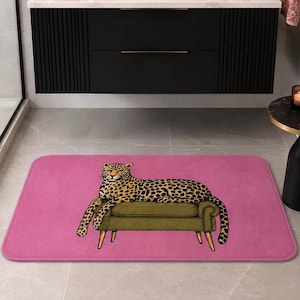 May include: A rectangular bath mat in a vibrant pink color featuring a central graphic illustration of a spotted leopard resting on a muted olive green upholstered bench with four light-colored wooden legs, centered on the floor in a bathroom.