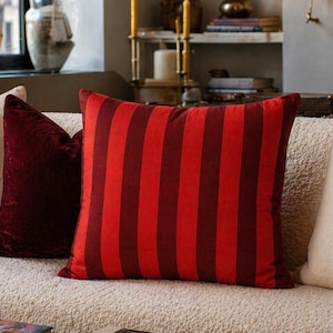 May include: A square throw pillow featuring vertical stripes in alternating shades of bright red and deep burgundy. The pillow is positioned on a textured cream-colored sofa, flanked by a solid dark red velvet pillow and a plain cream-colored pillow.