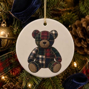May include: A round white ceramic ornament featuring a printed illustration of a teddy bear made of various dark-toned plaid fabric patches. The bear has a tan snout and paw pads, and the ornament hangs from a thin gold cord for display.