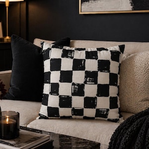 Könnte beinhalten: Ein dekoratives Zierkissen mit einem schwarz-weißen Schachbrettmuster liegt auf einem beigen Sofa. Weitere Kissen sind ein schwarzes Samtkissen und ein cremefarbenes strukturiertes Kissen. Eine brennende Kerze und Bücher liegen auf einem Marmor-Couchtisch.
