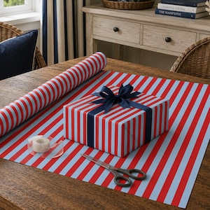 Puede incluir: Un rollo de papel de regalo y una caja cuadrada con un patrón de rayas verticales rojas y azul claro. La caja está envuelta con una cinta azul oscuro atada en un lazo. Se incluyen un rollo de cinta adhesiva transparente y unas tijeras de metal.