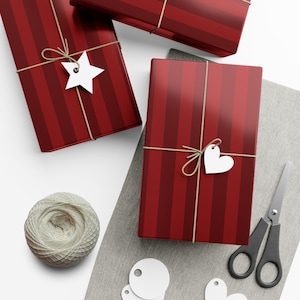 May include: Three wrapped gifts with red and burgundy vertical stripes, tied with twine and white tags. One tag is a star, another a heart. A ball of twine, scissors, and additional tags are also present.