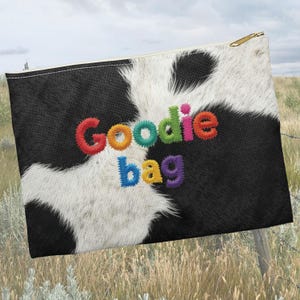 May include: A rectangular zippered pouch with a black and white cowhide pattern. The words "Goodie bag" are embroidered in colorful, rainbow-colored letters across the front. The zipper is gold-colored.