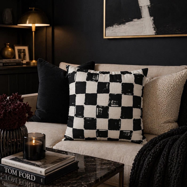 K&ouml;nnte beinhalten: Ein dekoratives Zierkissen mit einem schwarz-wei&szlig;en Schachbrettmuster liegt auf einem beigen Sofa. Weitere Kissen sind ein schwarzes Samtkissen und ein cremefarbenes strukturiertes Kissen. Eine brennende Kerze und B&uuml;cher liegen auf einem Marmor-Couchtisch.